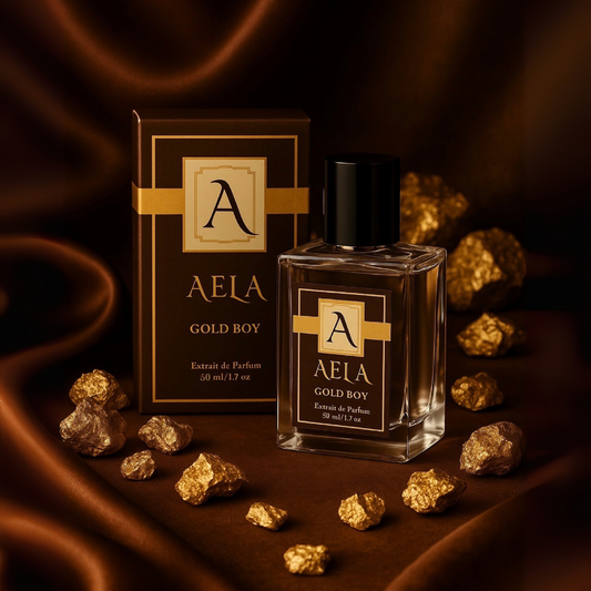 A clear rectangular perfume bottle with a black cap stands beside its matching box on a draped dark brown satin surface, surrounded by scattered gold crystals. The packaging and bottle label read AELA GOLD BOY.